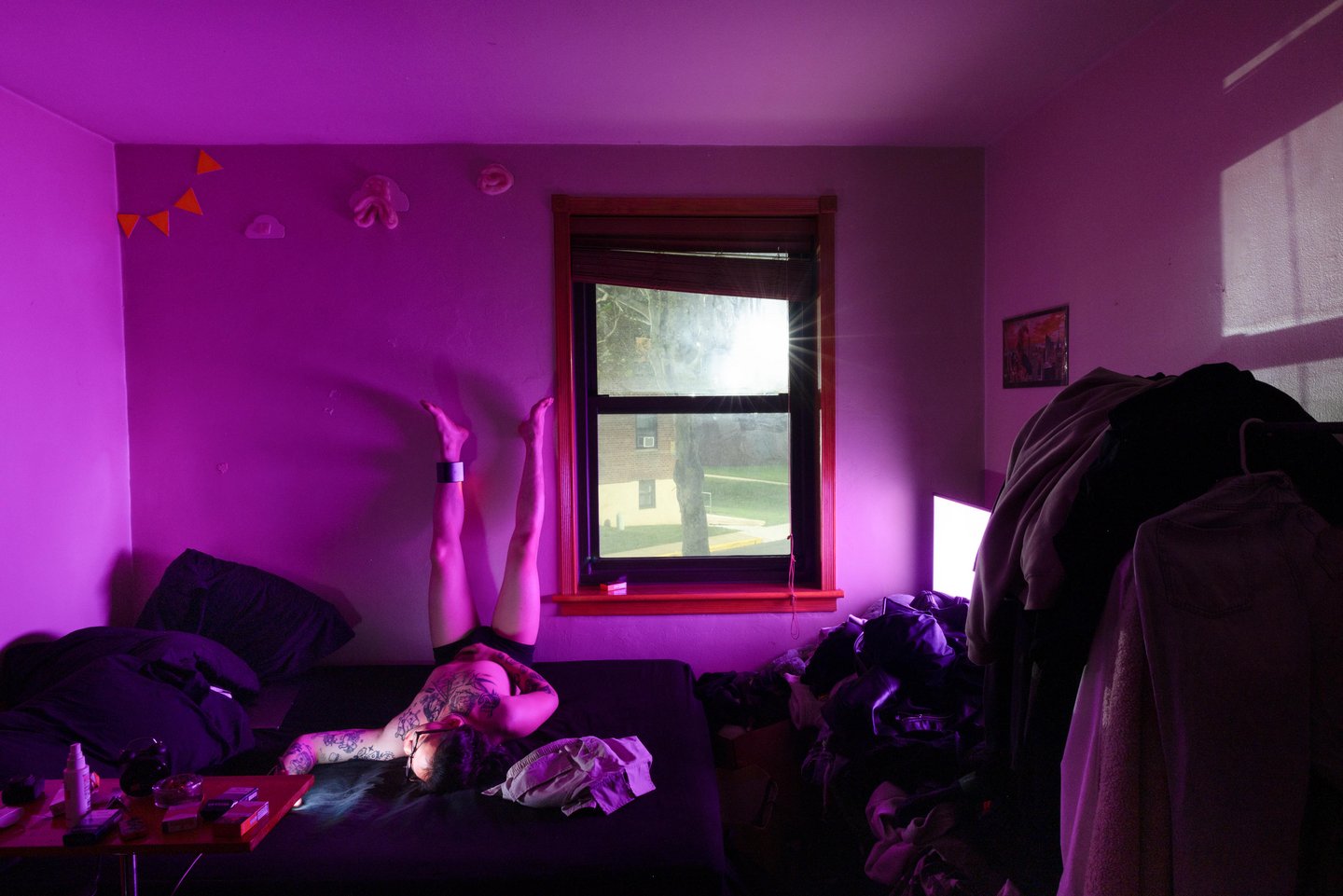 A bedroom washed in magenta light; a tattooed man lies on the bed with his legs up against the wall, sunlight bursting through the window — Win or Lose