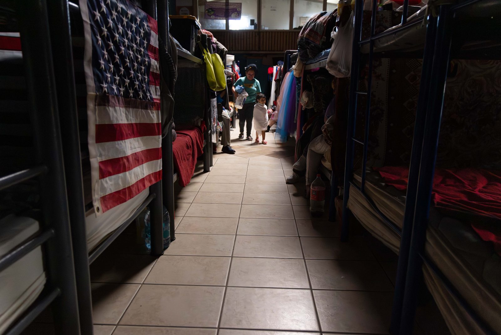 Inside a shelter dormitory, bunk beds line a long tiled corridor, an American flag hanging at the left, a woman and small child at the far end — The Liminal Innocence