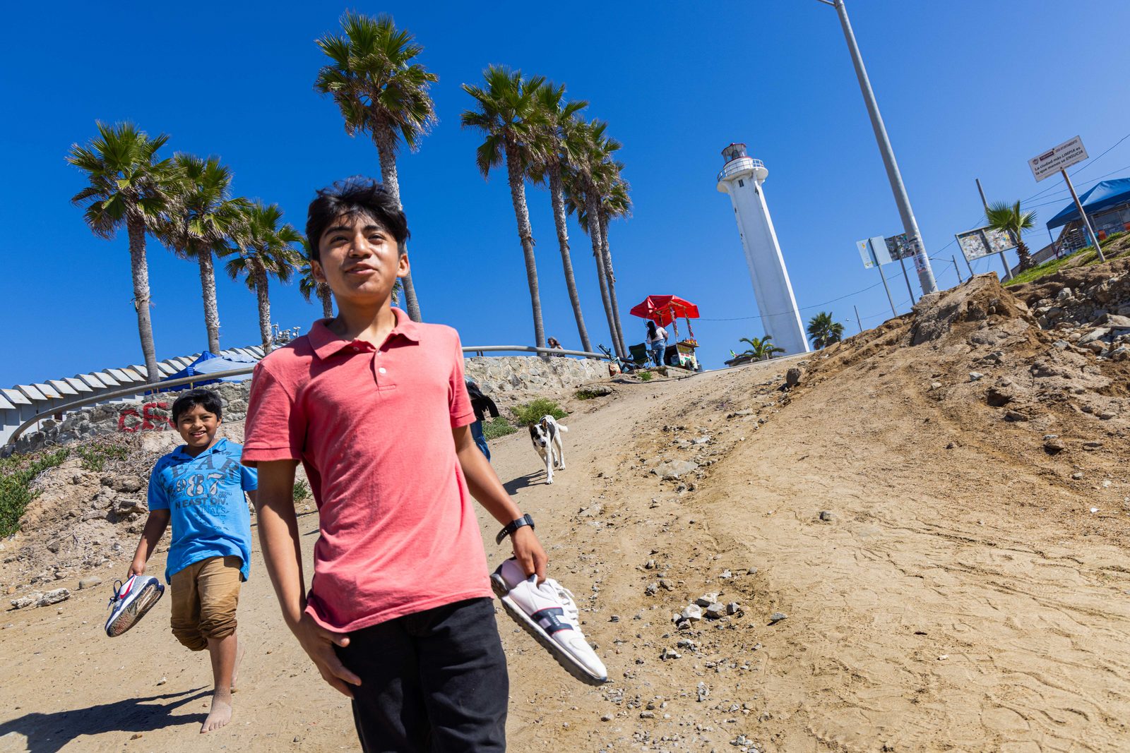 Two boys walking along a dusty path, palm trees and a lighthouse behind them — The Liminal Innocence