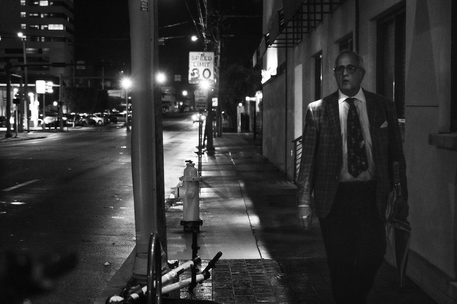 Black-and-white night street, a suited man walking past a fire hydrant beneath a speed-limit sign — Miami